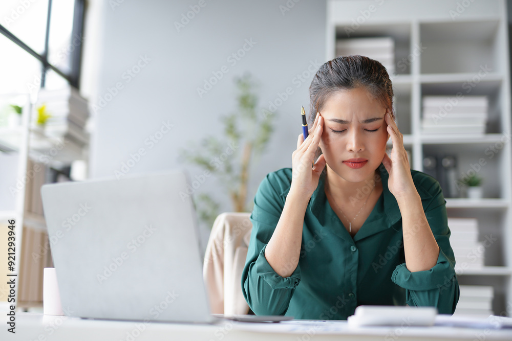Young businesswoman is feeling tired and having a headache, touching her head, while working on a laptop computer at the office Young businesswoman is feeling tired and having a headache, touching her head, while working on a laptop computer at the office