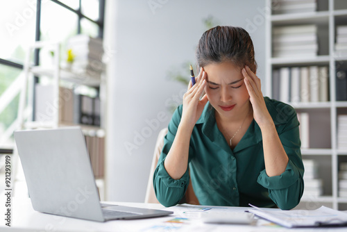 Stressed asian businesswoman touching her head, suffering from headache after working on laptop computer at office desk