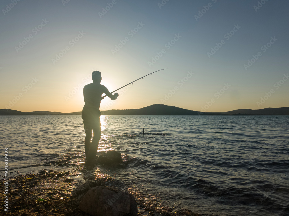 Fishing for pike, perch, carp. Fisherman with rod, spinning reel on sea or ocean. Man catching fish, pulling rod while fishing on sea, pond. Wild nature. The concept of rural getaway. Back view. Fishing for pike, perch, carp. Fisherman with rod, spinning reel on sea or ocean. Man catching fish, pulling rod while fishing on sea, pond. Wild nature. The concept of rural getaway. Back view.