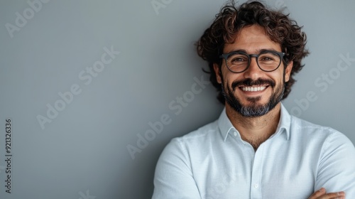 An amiable man with curly hair and glasses, dressed in a grey shirt and posing with arms crossed, radiates confidence and friendliness against a grey background, symbolizing trust and openness. photo