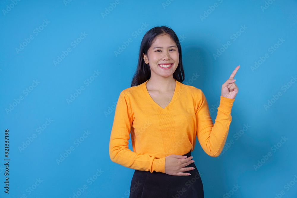 Fototapeta premium Smiling young woman wearing a yellow sweater is pointing her index finger at something on a blue background