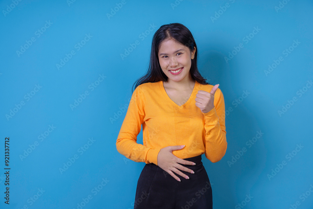 Young woman is smiling and showing a thumbs up gesture while touching her stomach on a blue background Young woman is smiling and showing a thumbs up gesture while touching her stomach on a blue background