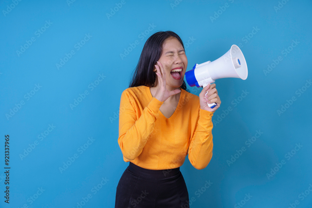 Young woman wearing an orange sweater is shouting loud in a megaphone in front of a blue background Young woman wearing an orange sweater is shouting loud in a megaphone in front of a blue background