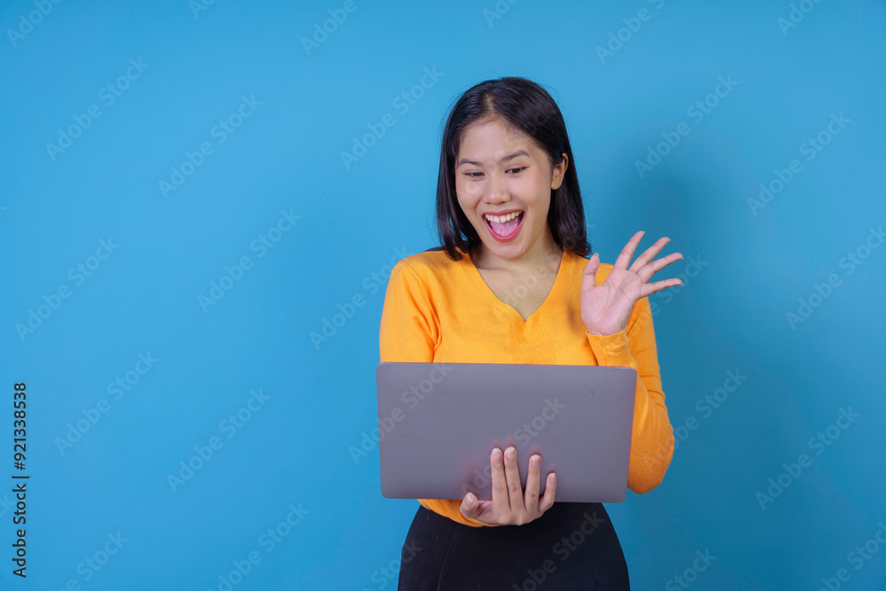 Excited young woman is waving and smiling while having a video call using a laptop on a blue background Excited young woman is waving and smiling while having a video call using a laptop on a blue background