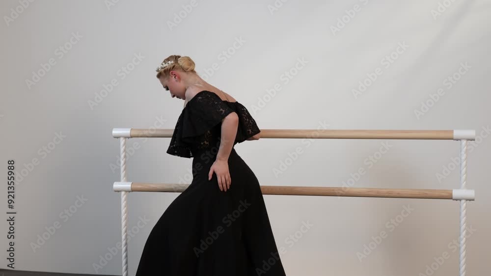 A beautiful ballerina in a black dress warming upi n a white sunny room with a black floor next to a ballet barre.
