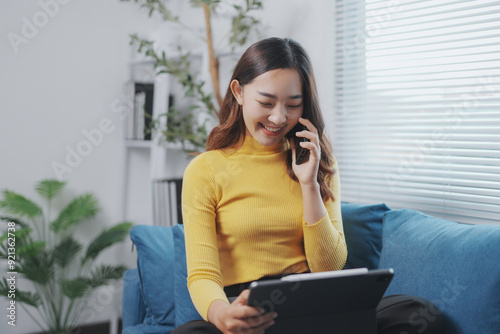 Young woman is working remotely, having a pleasant phone call while using her digital tablet, sitting comfortably on the sofa