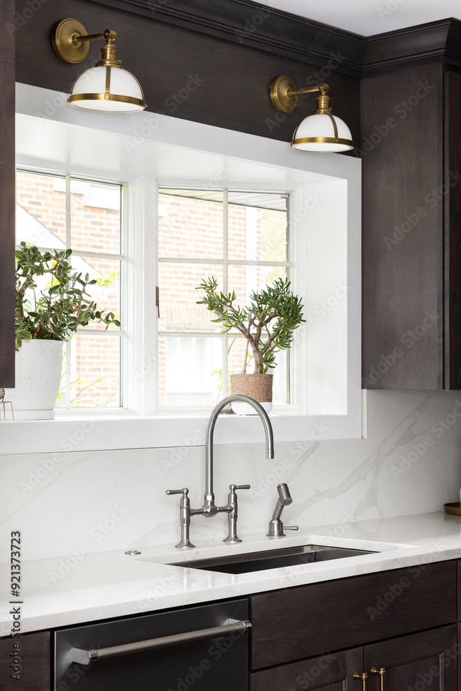 Φωτογραφία A kitchen faucet detail in a cozy kitchen with a stainless faucet, white marble countertop and backsplash, gold lights over a bright window, and dark wood cabinets