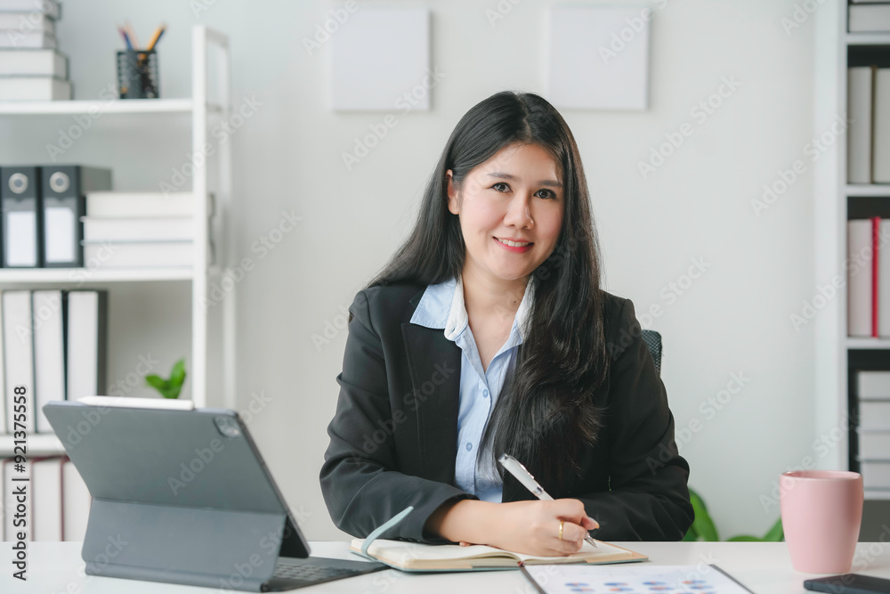 Asian businesswoman is sitting at her desk and taking notes, smiling towards the camera Asian businesswoman is sitting at her desk and taking notes, smiling towards the camera