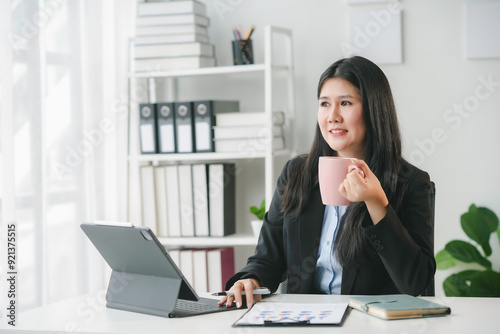 Businesswoman enjoying a cup of coffee while working on a digital tablet in her bright and modern office