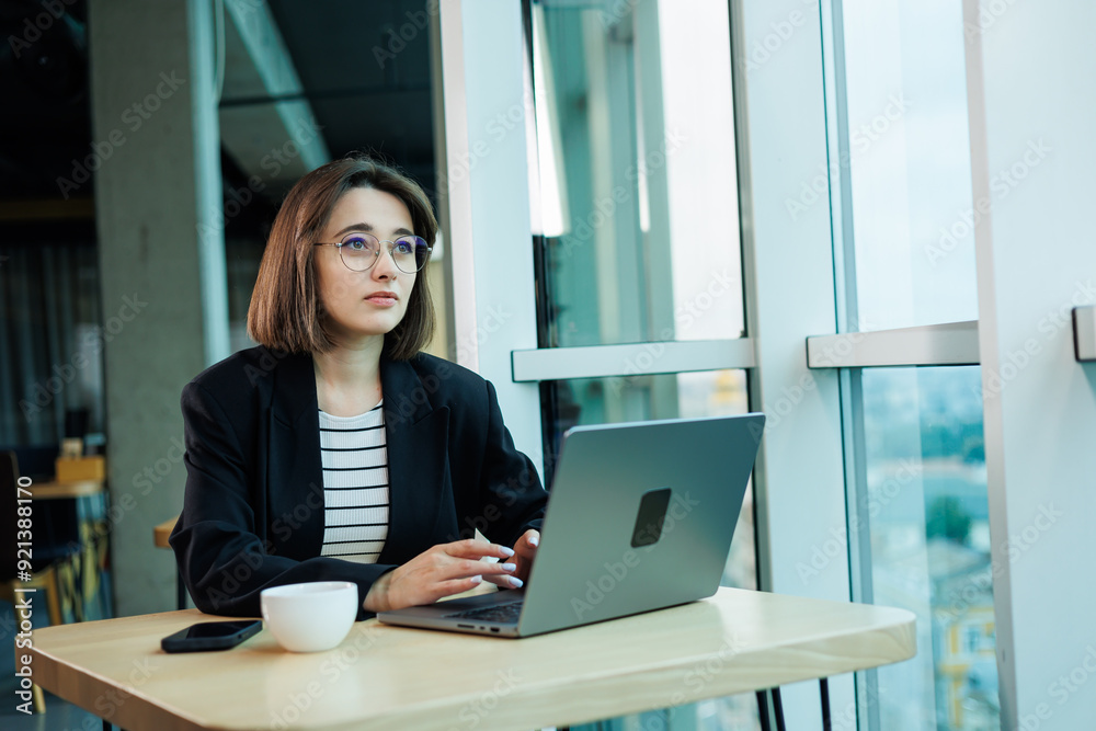 A young woman wearing glasses is sitting in a modern spacious working office. Woman office worker with laptop working. A young woman wearing glasses is sitting in a modern spacious working office. Woman office worker with laptop working.