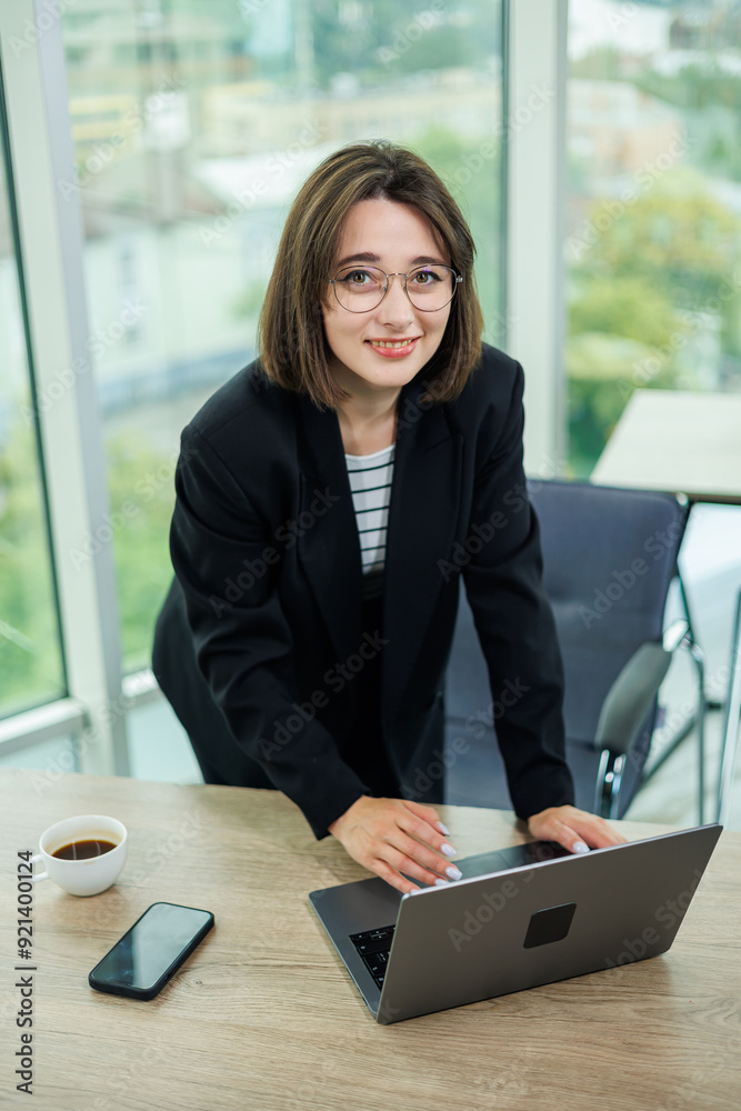 Woman business manager smiling working on laptop in office. An employee works in an office with large windows. Woman business manager smiling working on laptop in office. An employee works in an office with large windows.