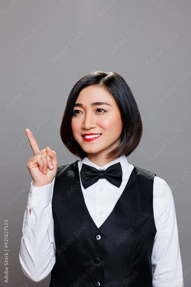 Cheerful asian woman wearing waitress uniform directing upwards with finger and posing in studio with joyful facial expression. Restaurant smiling receptionist pointing up Cheerful asian woman wearing waitress uniform directing upwards with finger and posing in studio with joyful facial expression. Restaurant smiling receptionist pointing up