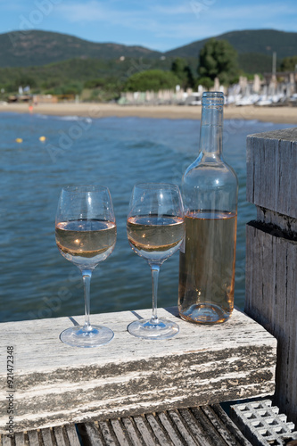 Bottle and glass of cold rose wine from Provence and wooden yacht boota pier on white sandy beach Plage de Pampelonne near Saint-Tropez, summer vacation in France photo