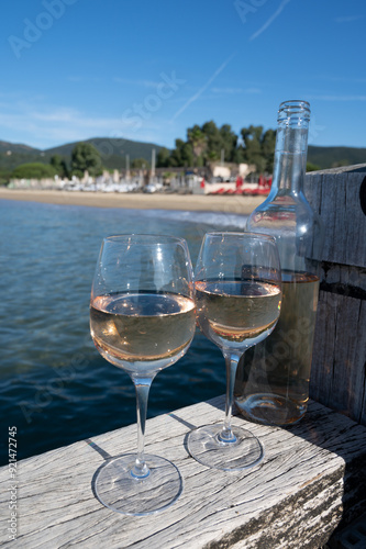 Bottle and glass of cold rose wine from Provence and wooden yacht boota pier on white sandy beach Plage de Pampelonne near Saint-Tropez, summer vacation in France photo