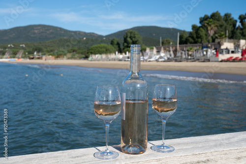 Bottle and glass of cold rose wine from Provence and wooden yacht boota pier on white sandy beach Plage de Pampelonne near Saint-Tropez, summer vacation in France photo