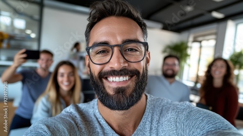 A bearded man smiling widely while taking a selfie with his friends indoors, capturing a joyful and memorable moment among a close group of friends.