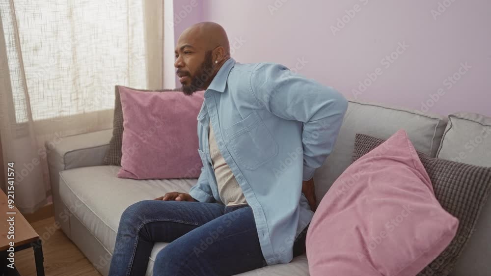Young man with beard experiencing back pain while sitting on a grey couch in a living room with soft lighting and pink cushions Young man with beard experiencing back pain while sitting on a grey couch in a living room with soft lighting and pink cushions