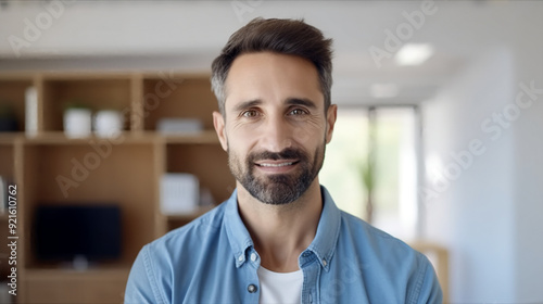 A portrait of an attractive businessman in his late thirties, with short hair and a beard wearing a blue shirt over a white t-shirt standing at the front facing the camera smiling.