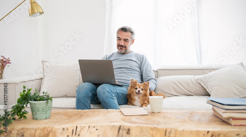 Portrait of senior old caucasian man stay home sit on the sofa  living room working on computer with lovely dog. Happy moment of pure love middle age male playing with little dog in winter