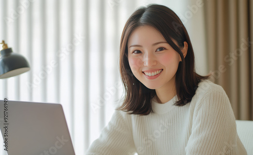 A young woman, smiling as she works on her laptop, is surrounded by a chic, minimalist home office interior in her beige shirt.