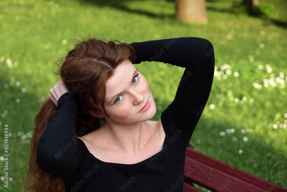 Attractive girl with long red hair and freckles fix her hairstyle sitting on a bench in summer park. Young sensual model wearing black top, female beauty