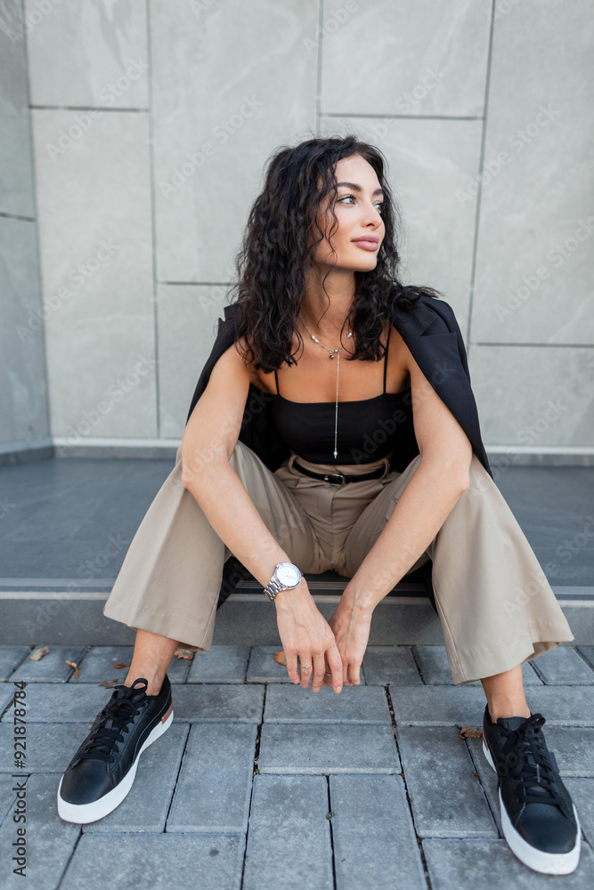 Pretty young beautiful woman with curly hair in elegant clothes with top, black blazer and pants with shoes sits in the city near a gray wall Pretty young beautiful woman with curly hair in elegant clothes with top, black blazer and pants with shoes sits in the city near a gray wall
