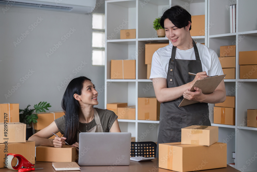 Happy Couple Managing Their Small Online Business from Home, Surrounded by Boxes and Working Together with a Laptop and Clipboard Happy Couple Managing Their Small Online Business from Home, Surrounded by Boxes and Working Together with a Laptop and Clipboard