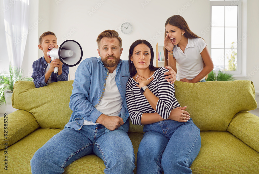 Tired exhausted family mother and father sitting on couch at home while noisy kids daughter and son shouting in loudspeaker in background. Problems of parenthood and need repose concept. Tired exhausted family mother and father sitting on couch at home while noisy kids daughter and son shouting in loudspeaker in background. Problems of parenthood and need repose concept.