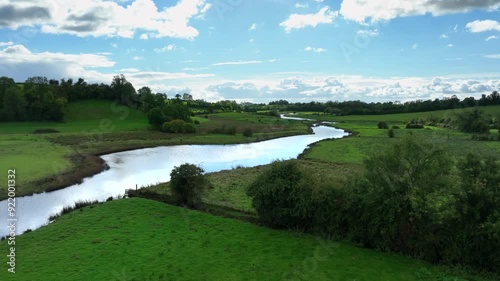 Finn River, County Monaghan, Ireland, September 2022. Drone pushes forward above trees growing on fence line with water blowing ripples over water reflecting cloudy sky as sheep graze in pasture. photo