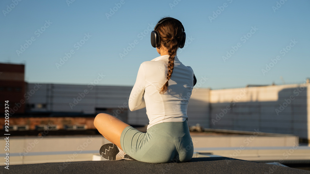 Fototapeta premium Young athletic girl sitting on concrete block on roof and meditating against buildings in city