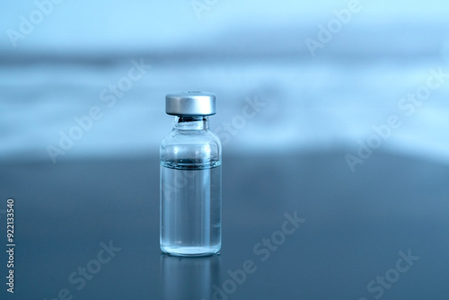 Transparent glass vial with an aluminum cap. The vial contains a clear liquid and rests on a reflective surface. The background is a soft, light blue blur