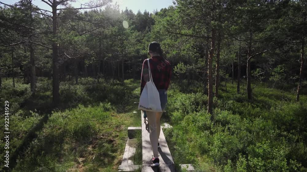 A woman in a red plaid shirt walks along a narrow wooden path in the middle of the thick Estonian forests. A young woman is accompanied by a small brown dog. A woman in a red plaid shirt walks along a narrow wooden path in the middle of the thick Estonian forests. A young woman is accompanied by a small brown dog.