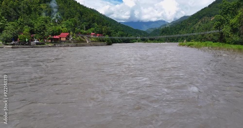 Drone shot of Bhotekoshi River flooding during monsoon in Nepal, with people living on the riverbank. Lush green hills, a bridge, and a beautiful sky after rain create a happy environment. photo