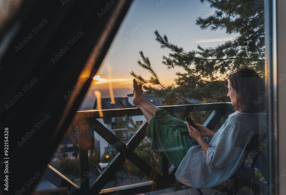 Young woman with smartphone greeting morning at dawn sitting on balcony overlooking mountains