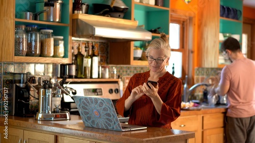 Smiling mature mother uses phone and works on laptop computer as teen son washes dishes as in kitchen.