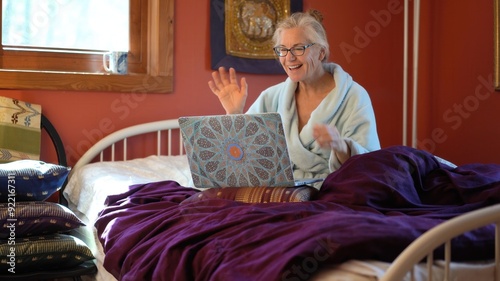 Smiling happy mature woman talks on a teleconference on her laptop computer while wearing a bathrobe in bed.