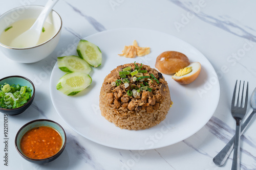 A white plate of Thai-style rice topped with minced chicken, served with sliced cucumber, a halved boiled egg, green onions, fried garlic, and chili sauce, alongside a soup bowl,