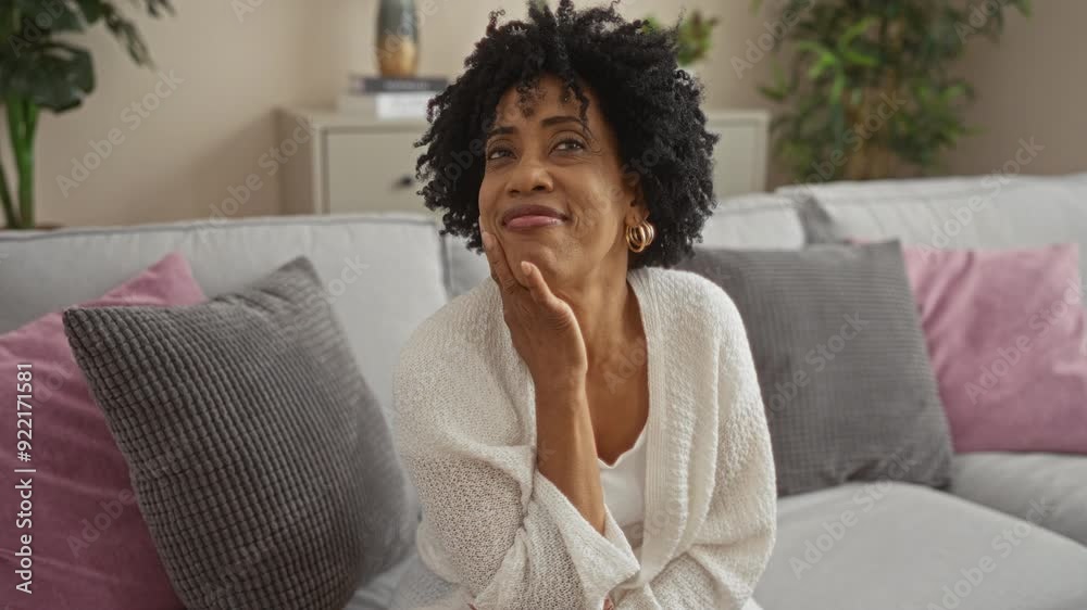 A beautiful african american woman with curly hair smiling in a cozy living room with cushions on a sofa. A beautiful african american woman with curly hair smiling in a cozy living room with cushions on a sofa.