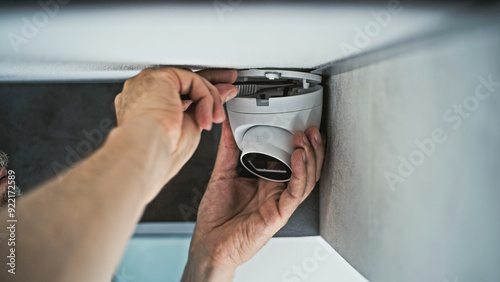 Professional male installer screws CCTV camera to the ceiling in office room using screwdriver tool and adjusts the angle. Monitoring and tracking. Video surveillance, security system, and privacy. photo