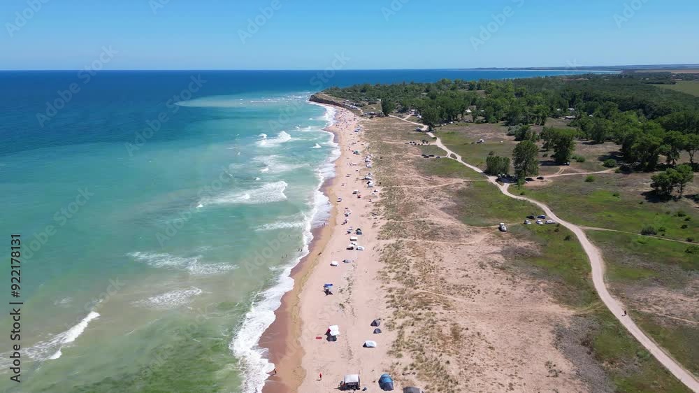 Aerial view of a sandy beach with scattered tents and umbrellas along the shoreline. Waves gently wash onto the shore, capturing a peaceful and relaxing summer day by the ocean Aerial view of a sandy beach with scattered tents and umbrellas along the shoreline. Waves gently wash onto the shore, capturing a peaceful and relaxing summer day by the ocean