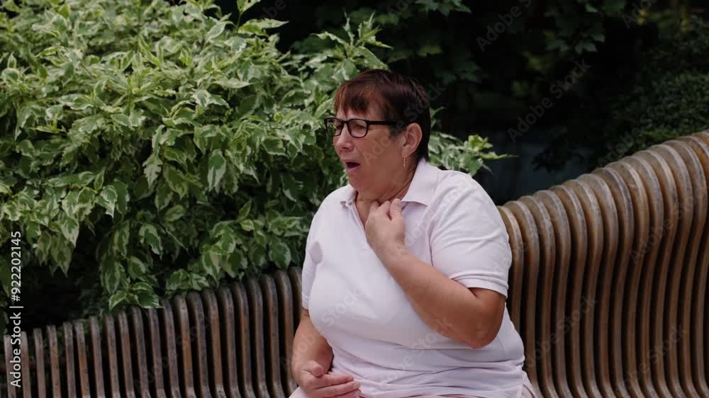 Elderly woman wearing pink polo shirt and glasses is sitting on modern wooden bench. Grandmother feels a sore throat and is coughing. In the background, there is greenery. Elderly woman wearing pink polo shirt and glasses is sitting on modern wooden bench. Grandmother feels a sore throat and is coughing. In the background, there is greenery.