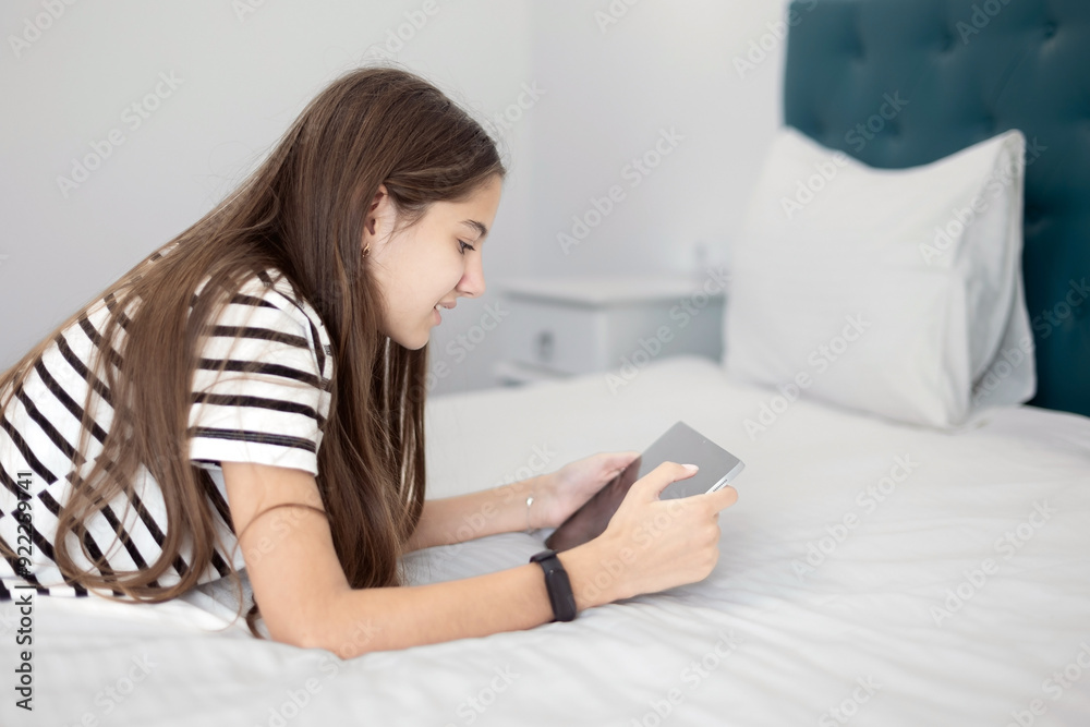 Teenage Girl Using Tablet in a Comfortable Bedroom. A teenage girl with long brown hair lying on a bed while using a tablet. Teenage Girl Using Tablet in a Comfortable Bedroom. A teenage girl with long brown hair lying on a bed while using a tablet.