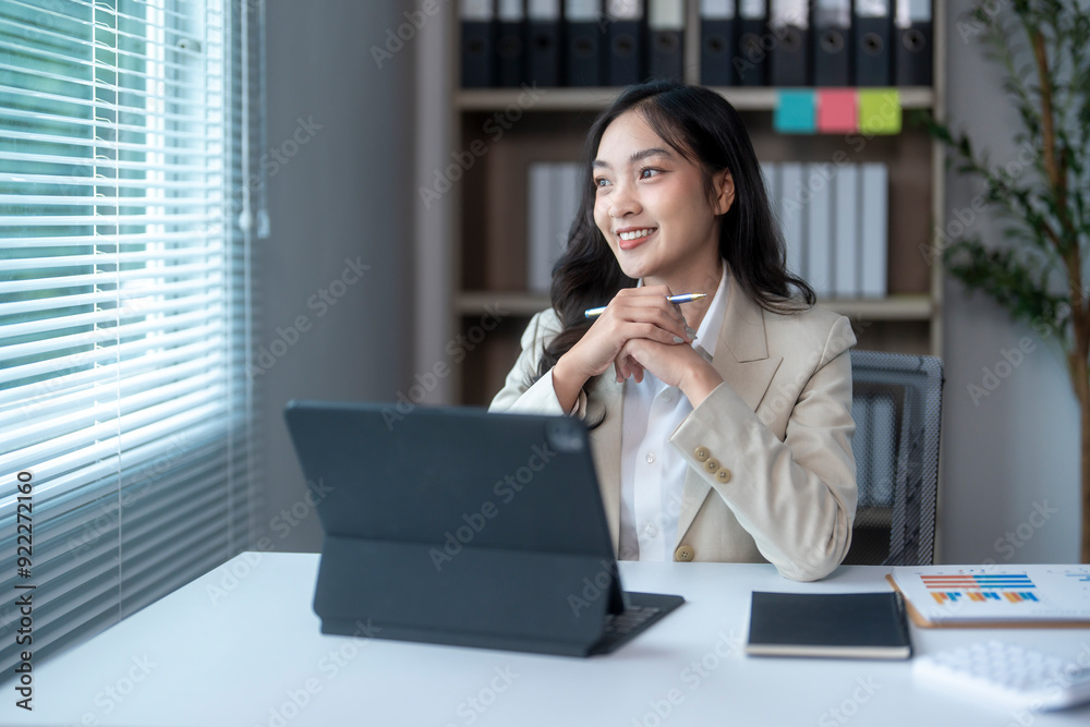 Businesswoman smiling and looking out the window while working on a tablet in her office Businesswoman smiling and looking out the window while working on a tablet in her office