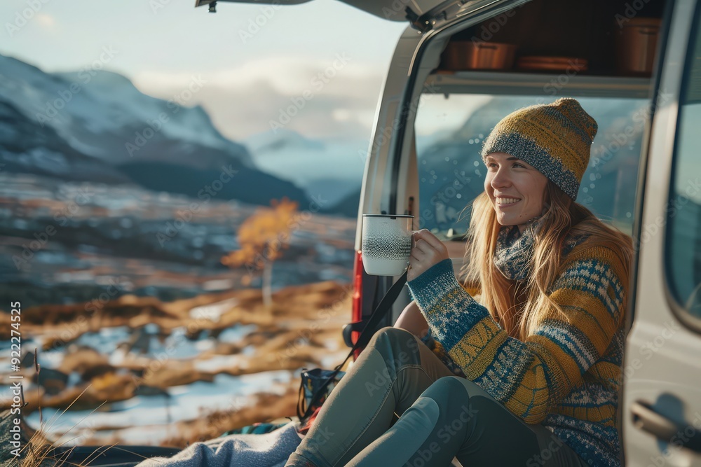 Naklejka premium Van life: Woman greeting sunrise from camper van trunk, sipping hot tea or coffee, bundled in warm sweater with snowy mountains in the background.
