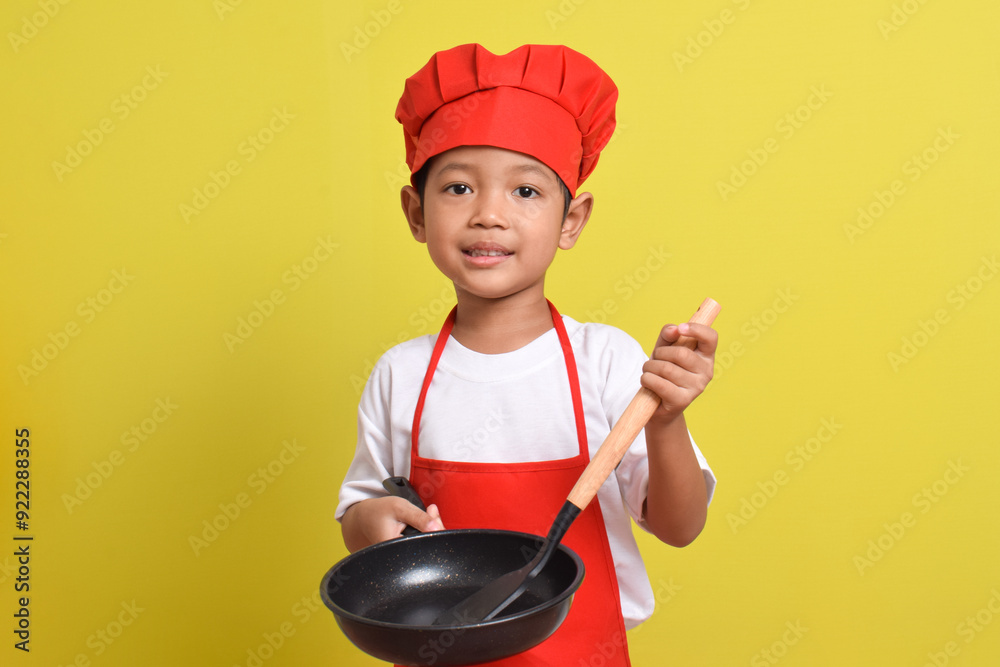 Cute little chef holding frying pan and spatula. Asian boy wearing red apron and hat on isolated yellow background Cute little chef holding frying pan and spatula. Asian boy wearing red apron and hat on isolated yellow background