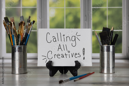 Calling all creatives! sign sitting on easel in front of a bay window with different sizes of paint brushes.