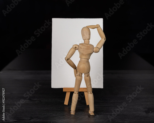 Blank white sign sitting on art easel with wooden mannequin standing in front of empty canvas scratching head. photo