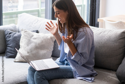 Young woman sitting on a couch in her living room, excited and surprised while using a computer for an online class, happily interacting and gesturing with friends or a partner on screen