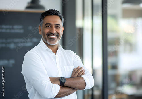 Portrait of a happy middle-eastern man in a white shirt, standing in an office setting, smiling with his arms crossed and looking directly at the camera