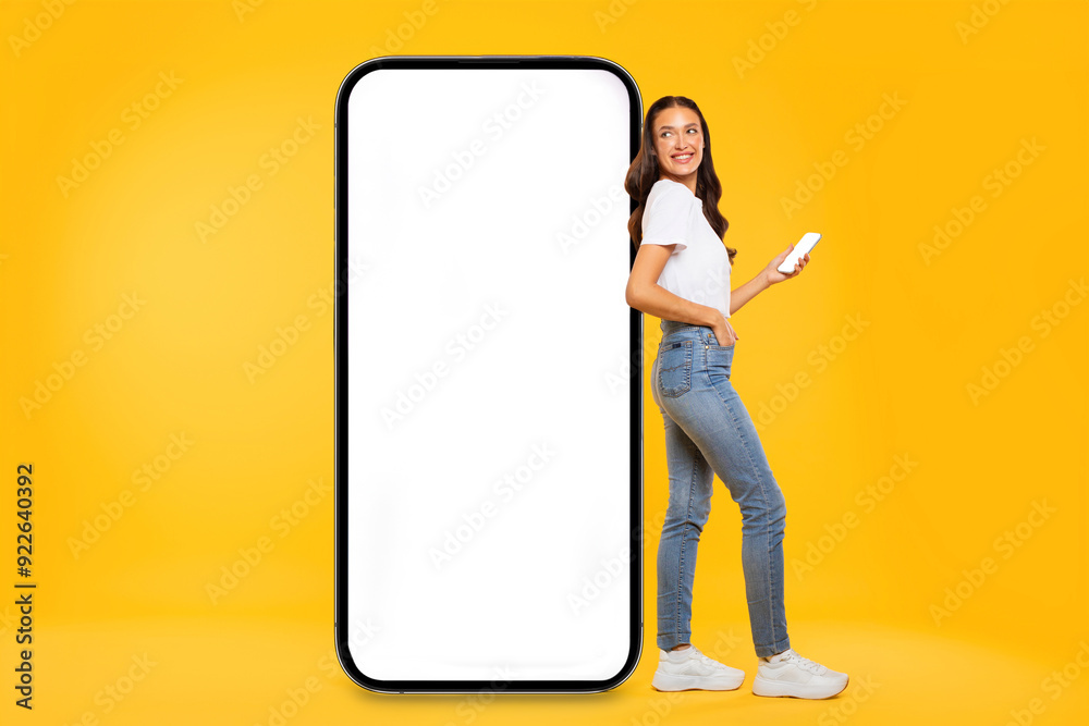 Happy young European lady standing by huge smartphone with white blank screen, showing online offer, yellow studio background, mockup Happy young European lady standing by huge smartphone with white blank screen, showing online offer, yellow studio background, mockup